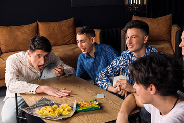 Five friends spending time together, having fun, playing table games and eating fast food and snacks. Small group of friend in a living room. man losing and raging at his friends. - obrazy, fototapety, plakaty