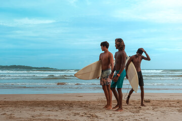 company of young guys Sri Lankans, Indians surfers on the ocean coast with a surfboard, sporty tanned body, smiling and running with a surfboard on the beach on a sunny summer day, ready to surf.