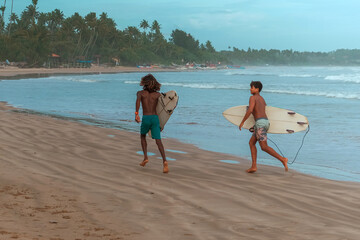 Obraz premium company of young guys Sri Lankans, Indians surfers on the ocean coast with a surfboard, sporty tanned body, smiling and running with a surfboard on the beach on a sunny summer day, ready to surf.