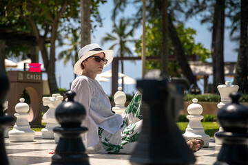 A beautiful girl is playing giant chess. Sunlight. Chessmen. Girl in a hat and sunglasses.
