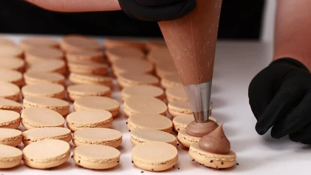 Making french desert macarons. Chef hands with confectionery bag add cream to the ready halves of brown chocolate macaroons at pastry shop. cooking, food and baking concept