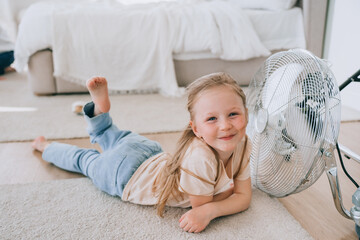 Pretty cheerful little girl in casual laying on floor at bedroom close to fan at hot summer day...