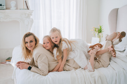 Succesful businesswoman in beige suit laying on bed at home with two little daughters looks at camera happy to be with family. Domestic leisure, motherhood. Maternity, childhood, women.