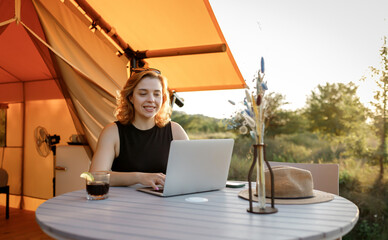 Smiling Woman freelancer using a laptop on a cozy glamping tent in a sunny day. Luxury camping tent...