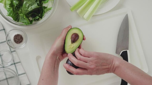 Woman cuts avocado on a white cutting board, view from above. Celery lettuce salad with avocado and lemon dressing recipe