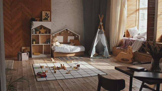 Zoom in shot of spacious nursery room with cozy interior and no people during day. There is teepee, baby bed, carpet with toys, shelves with home decor, pillows and plaid on window sill