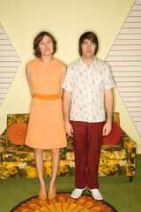 Caucasian mid-adult couple wearing retro clothes standing stiffly in room decorated with vintage furniture.