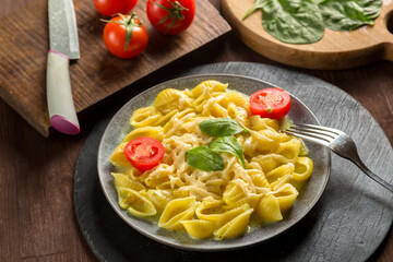 Pasta in cream sauce with cheese garnished with spinach with a fork on a wooden stand next to tomatoes, a knife and greens.