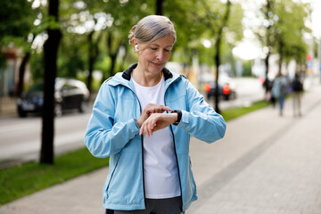 Sporty Mature Runner Lady Standing in the Street, Listening to Music on the Earphones, Wearing a Smartwatch, and Using it to Watch Videos. Sport and Healthy Lifestyle Concept