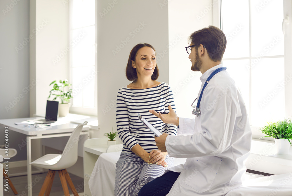 Wall mural happy middle-aged female patient talking to her family doctor in hospital office. woman listens to a