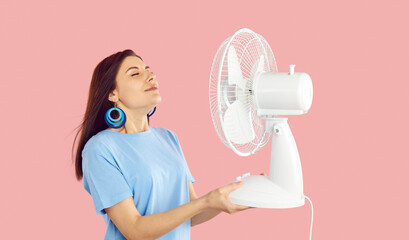 Overheated millennial girl isolated on pink studio background hold fan breathe fresh air. Unwell thirsty young woman use ventilator suffer from hot summer weather. Heatstroke and ventilation. - obrazy, fototapety, plakaty