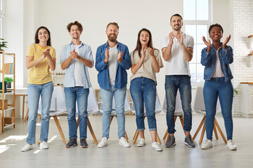 Obraz premium Friendly multiracial people in casual clothes stand in row and applaud looking at you. Group of friends in jeans and T-shirts joyfully clap their hands while standing in row in room. Team concept.