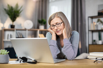 Exhausted middle-aged woman in glasses feeling tired while looking at laptop screen after full day of work in remote office. Senior lady being overcome with fatigue after online brainstorm session.