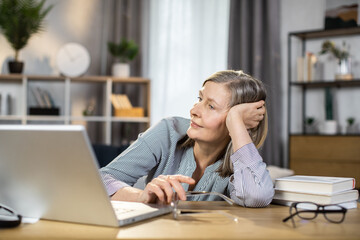 Exhausted middle-aged woman in glasses feeling tired while looking at laptop screen after full day of work in remote office. Senior lady being overcome with fatigue after online brainstorm session.