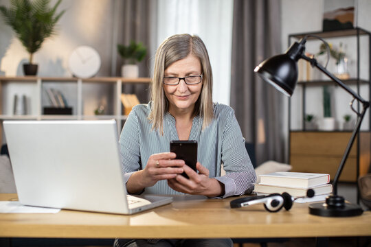 Mindful senior woman in spectacles looking at phone screen while operating modern laptop at office desk in home interior. Efficient freelancer checking meetings schedule via mobile application.