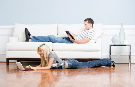 Man reads on a couch while woman stretches out on the floor with her laptop. Horizontal format.