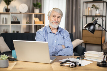 Portrait of senior caucasian man posing with his arms crossed while sitting at office desk with computer and headset. Friendly employee taking break from remote work while staying in cozy home office.