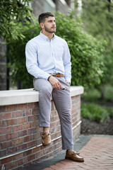 Fototapeta premium Close up portrait of bearded young man wearing light blue button up shirt sitting on retaining wall in a park