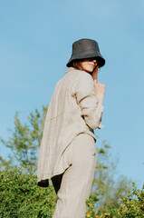 Young woman in linen outfit and bucket hat posing on a hill with sky as background