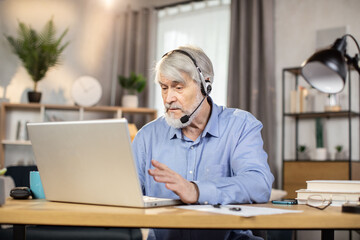 Serious middle-aged male in headset with microphone making video call while sitting at writing desk...