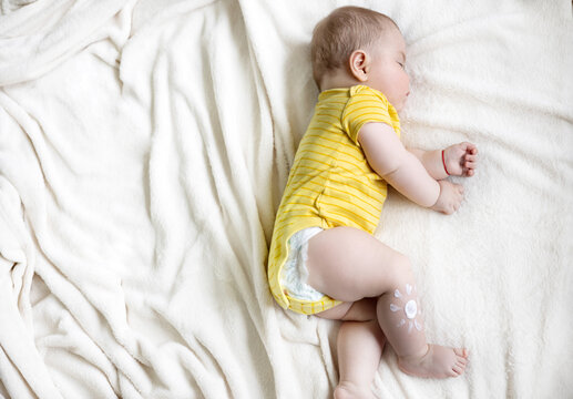sleeping adorable baby boy on blanket with drawn sun painted from body lotion on leg.mother hand applying cream sun protection sunblock.vacation.fat toddler infant cute feet child in yellow bodysuit