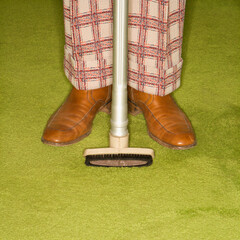 Close-up of Caucasian male legs in plaid pants with vacuum extension standing on green retro carpet.
