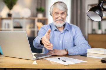 Selective focus on male hand extended for handshake by elderly man in headset and glasses sitting at writing desk indoors. Friendly remote employee greeting warmly new client at distant workplace.