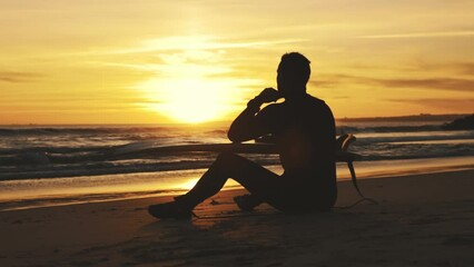 Silhouette of a thoughtful man surfer sitting on the seashore at sunset