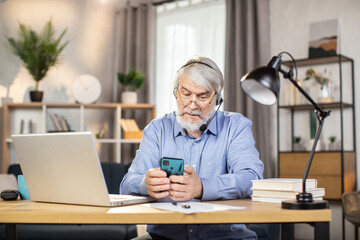 Fototapeta na wymiar Focused elderly adult in headset and glasses holding smartphone while sitting at desk with portable computer on it. Efficient employee texting short message in online chat via app in home office.