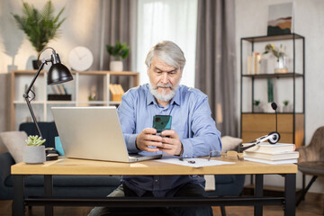 Focused elderly adult in headset and glasses holding smartphone while sitting at desk with portable computer on it. Efficient employee texting short message in online chat via app in home office.
