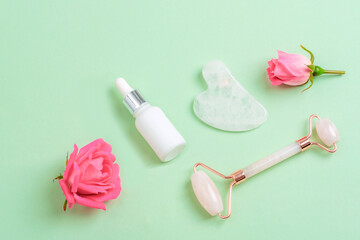 White cosmetic serum bottle, face roller, gua sha stone and pink rose flowers on light green background. Skin care, beauty treatment concept. Top view, flat lay, copy space