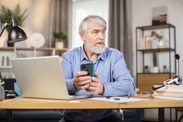 Fototapeta premium Focused elderly adult in headset and glasses holding smartphone while sitting at desk with portable computer on it. Efficient employee texting short message in online chat via app in home office.
