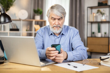 Mindful caucasian man of mature age holding cell phone while teleworking in home interior. Professional business leader looking through contact information on smart gadget at desk in remote office.