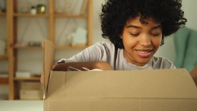 African girl unpacking delivery looking in box. Happy woman opening carton box. Female getting parcel looking at delivered goods items. Satisfied client positive feedback quick express deliver