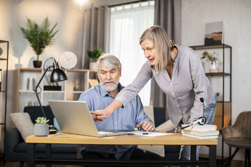 Middle-aged woman in business casual wear pointing at laptop screen while senior man sitting at writing desk in home office. Focused partners analyzing data of digital report in remote workplace.
