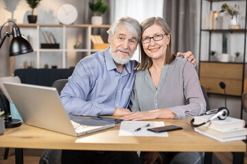 Elegant senior lady in glasses and gray-haired elderly man utilizing portable computer for distant work at home. Efficient freelancers studying details of temporary contract on digital screen.