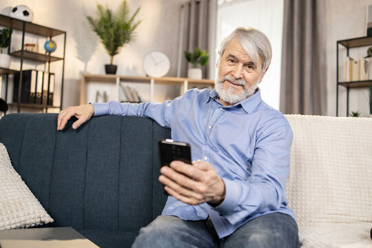 Focus on senior caucasian male in casual clothes posing with smartphone while taking seat on dark sofa in living room. Relaxed gray-haired man using mobile connection for calls and messages.