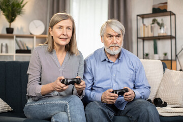 Mindful couple of senior people sitting on sofa with black input devices and looking attentively on screen in room interior. Elderly adults entertaining by playing video games using controllers.
