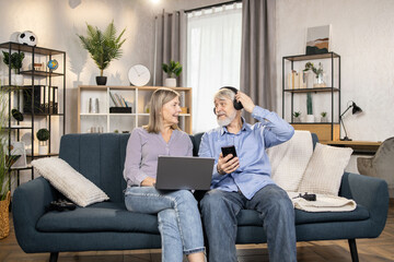 Cheerful mature man using wireless headphones and mobile while beautiful middle-aged woman relaxing with laptop in lounge. Senior spouses listening to excellent music from modern playlist at home.