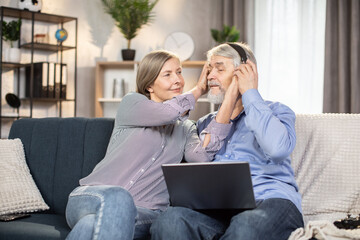 Lovely elder married couple resting on couch of beautiful apartment and using personal laptop for watching romantic movies. Caring wife helping husband to put on wireless headphones.