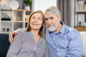 Lovely senior couple relaxing on sofa in living room enjoying retirement and free time at home. Loving husband and wife in casual clothes posing together at camera. Concept of happiness in marriage.