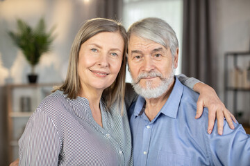 Portrait of devoted couple standing close together, embracing and looking straight at camera. Mature woman holding hand gently on shoulder of grey-haired husband. Concept of marital love and care.