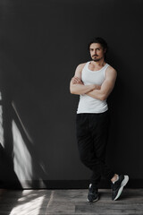 Fototapeta na wymiar Self assured muscular bearded man with black hair in white sleeveless t shirt and pants standing on black background and looking at camera