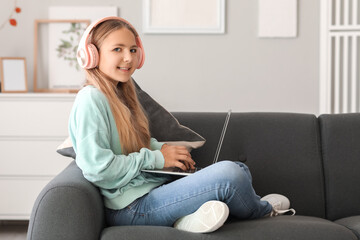 Little girl in headphones using laptop on sofa at home