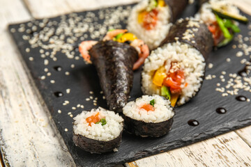 Slate board with tasty sushi rolls, cones, sauce and sesame seeds on light wooden background, closeup