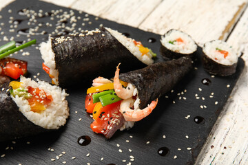 Slate board with tasty sushi rolls, cone, sauce and sesame seeds on light wooden background, closeup