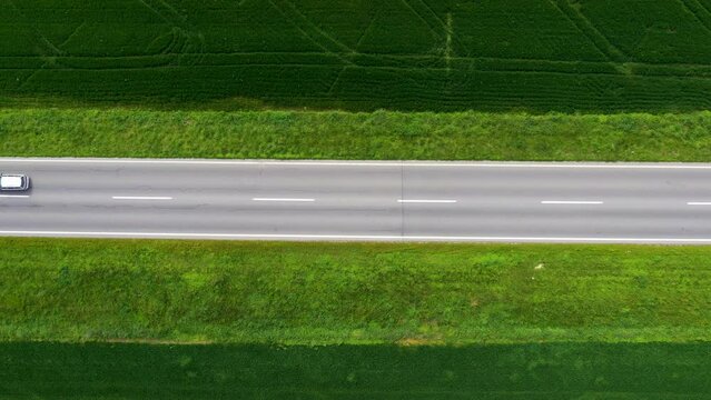 Top view drone pov aerial shot of white car driving along a straight road through cultivated landscape