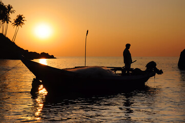 Silhouette of fisherman with nets in the sea on his boat at the dusk. Palm trees on the hill on background