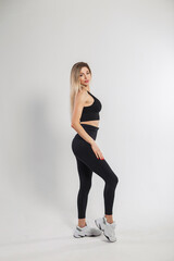 Beautiful young blonde fitness girl in fashionable black sportswear with sneakers, leggings and a top on a white background in the studio