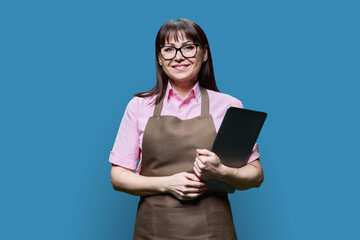 Portrait of middle aged woman in apron with laptop on blue background